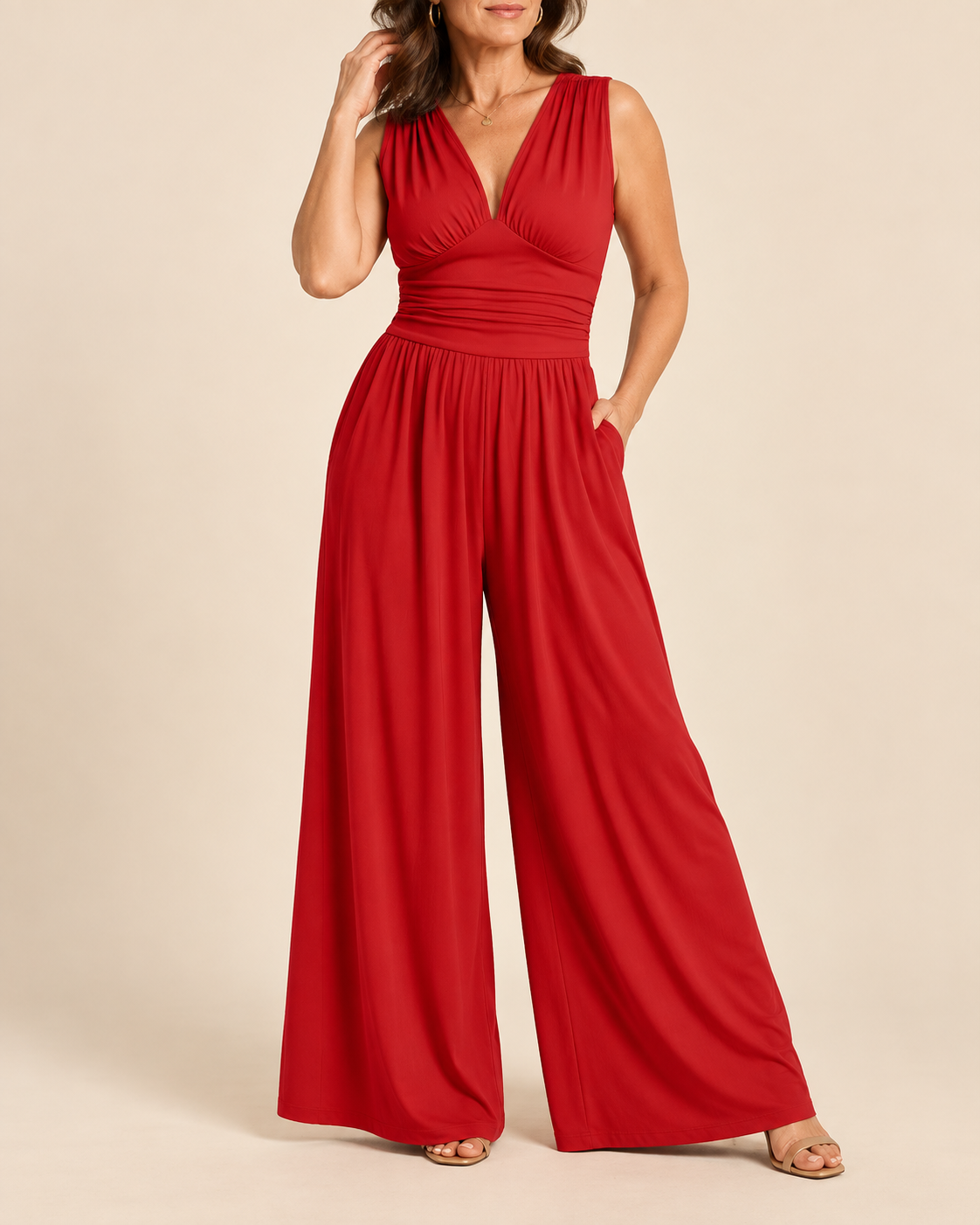 A woman modeling a bright red sleeveless jumpsuit with a draped V-neck bodice, gathered waist, and wide legs, photographed against a plain beige studio background.