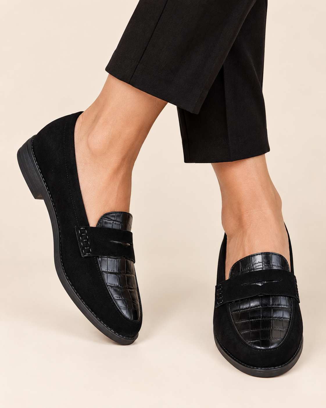 A pair of black loafers worn with black cropped trousers, shown on a model's feet against a light beige studio background. The loafers feature a mix of matte suede and crocodile-embossed leather panels.