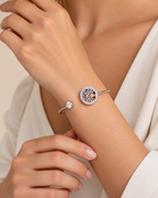 A close-up of a model's wrist wearing two silver-toned bracelets: one with a round tree-of-life motif surrounded by small clear stones, and another with a single round clear stone, against a soft cream-colored top.