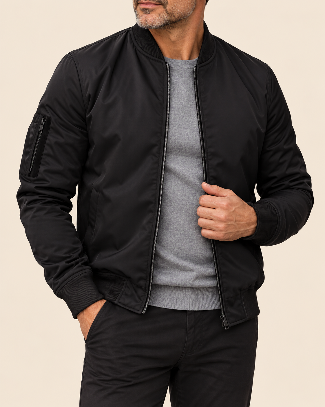 A man wearing a black bomber jacket over a gray crewneck sweater and black pants, shown from the chin down against a plain beige background.
