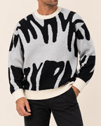 A man wearing a black and off-white zebra-patterned knit sweater with cream ribbed collar, cuffs, and hem, paired with black pants, against a solid light beige background.