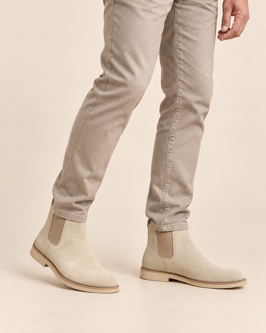 Beige suede Chelsea boots with elastic side panels, shown on a model wearing slim-fit beige jeans against a solid light beige background.