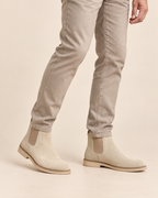 Beige suede Chelsea boots with elastic side panels, shown on a model wearing slim-fit beige jeans against a solid light beige background.