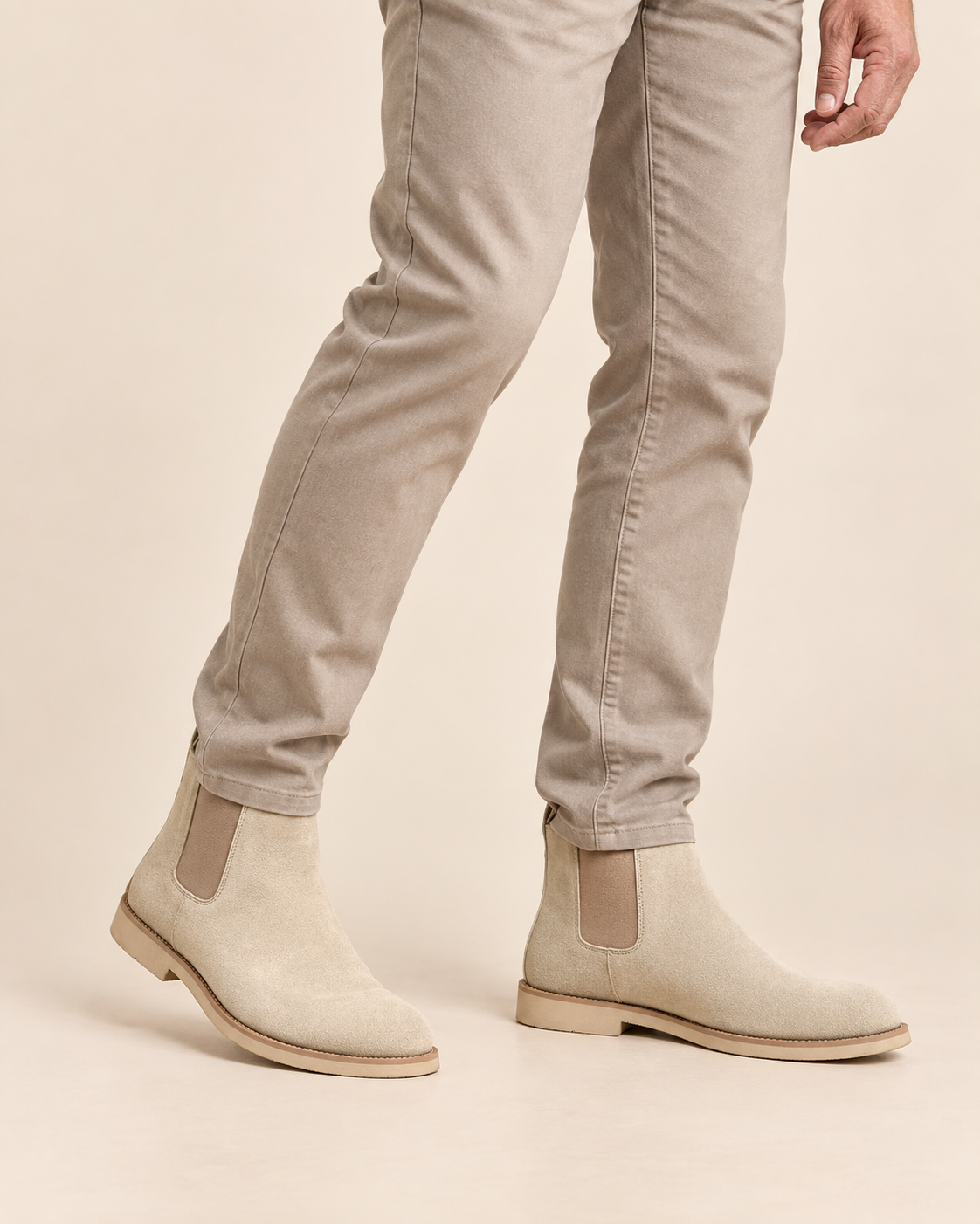 Beige suede Chelsea boots with elastic side panels, shown on a model wearing slim-fit beige jeans against a solid light beige background.