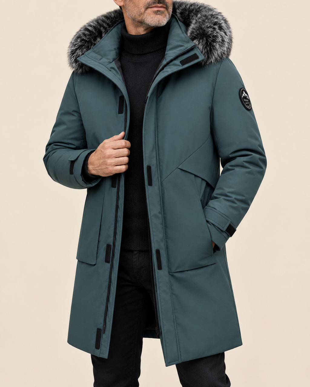 A man wearing a mid-length teal parka with a gray faux-fur trimmed hood, black turtleneck, and black pants, photographed against a plain beige studio background.
