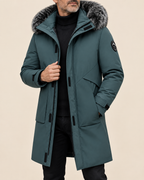 A man wearing a mid-length teal parka with a gray faux-fur trimmed hood, black turtleneck, and black pants, photographed against a plain beige studio background.