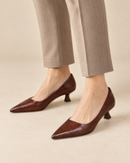 A person wearing pointed-toe brown pumps with kitten heels, paired with beige trousers, shown against a warm beige studio background.