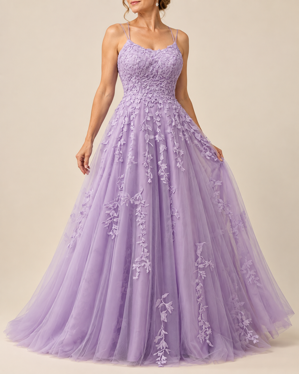 A woman wearing a lavender purple A-line formal gown with floral lace appliques and thin spaghetti straps, photographed against a solid beige studio background.