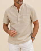 A man wearing a light beige linen short-sleeve henley shirt with a chest pocket, paired with white pants, against a solid beige background.