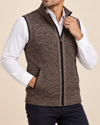 A man wearing a brown and black textured zip-up knit vest over a white collared shirt and dark pants, shown from the neck down against a plain beige studio background.