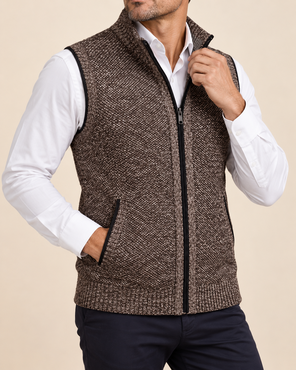 A man wearing a brown and black textured zip-up knit vest over a white collared shirt and dark pants, shown from the neck down against a plain beige studio background.