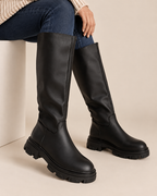 A pair of black knee-high leather boots with chunky lug soles, shown on a model wearing blue jeans against a warm beige background.