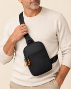A man wearing a cream-colored knit sweater and khaki pants models a black crossbody sling bag with an orange zipper pull against a solid beige background.