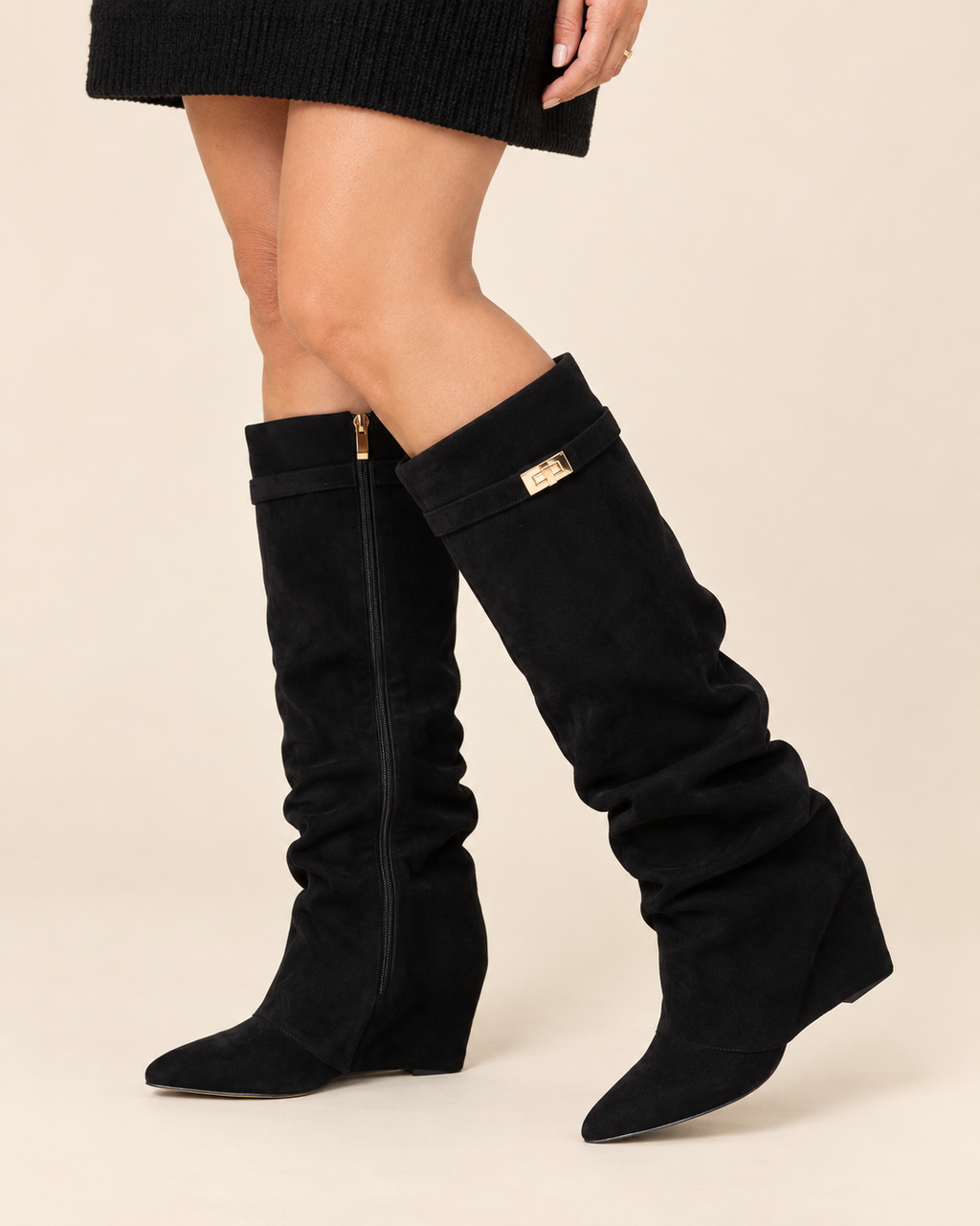 A pair of black knee-high slouchy suede boots with wedge heels, shown on a model against a light beige background. One boot features a side zipper with a gold pull tab, and the other has a small gold rectangular hardware accent.