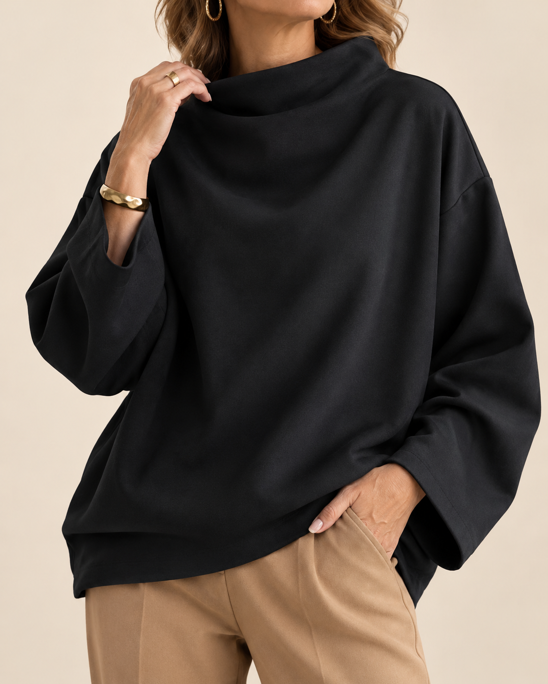 A woman wearing a black oversized long-sleeve top with a wide draped neckline, paired with tan pants, against a plain beige background.