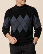 A man wearing a black mock-neck knit sweater with a dark gray argyle pattern on the chest and sleeves, paired with black trousers, against a solid beige background.
