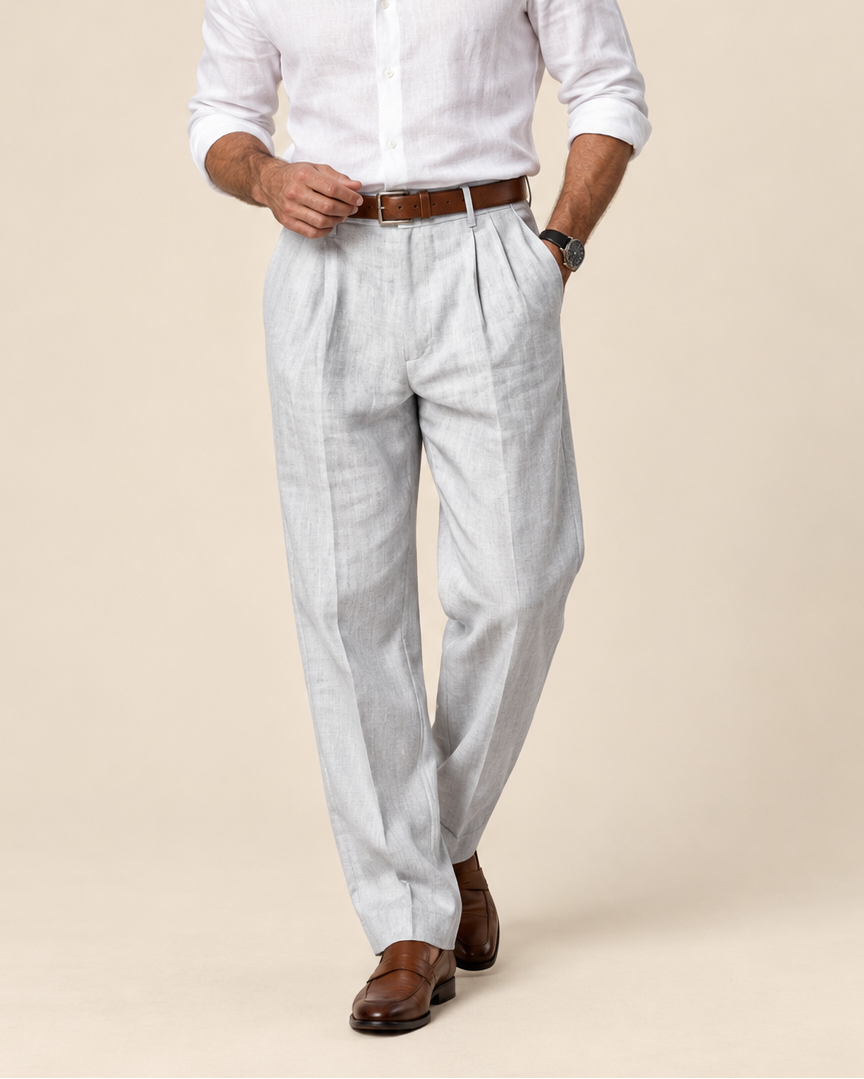 A man modeling a white long-sleeve linen shirt and light gray pleated linen trousers with a brown leather belt, styled with brown leather dress shoes against a solid beige studio background.