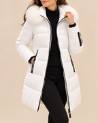A woman wearing a long white puffer coat with a large fluffy white fur-trimmed hood, black zipper details, and black side accents, paired with a black turtleneck and black pants, against a solid beige background.