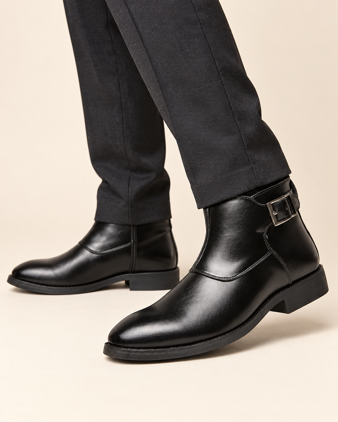Black leather ankle boots with a side buckle strap, worn with dark gray trousers on a light beige background.