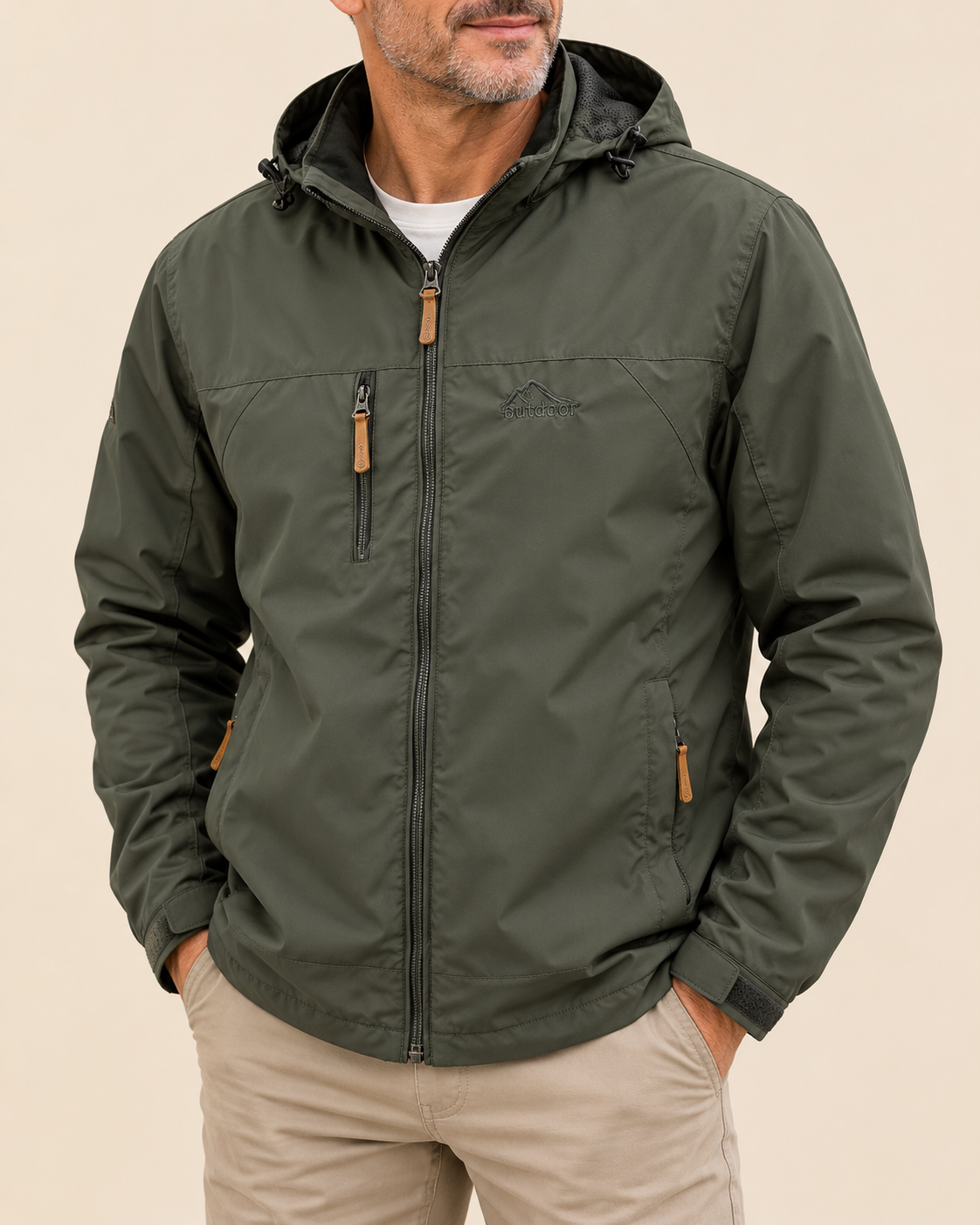 A man wearing an olive green hooded jacket with a full front zipper, zippered chest pockets, and tan zipper pulls, paired with beige pants against a plain beige background.