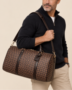 A man wearing a black quarter-zip sweater and beige pants carries a brown and tan checkered duffel bag with dark brown leather straps and gold-tone hardware against a light beige studio background.