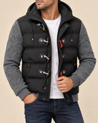 A man wearing a black hooded puffer vest with gray quilted sleeves, toggle closures, and a white t-shirt underneath, paired with blue jeans against a beige background.