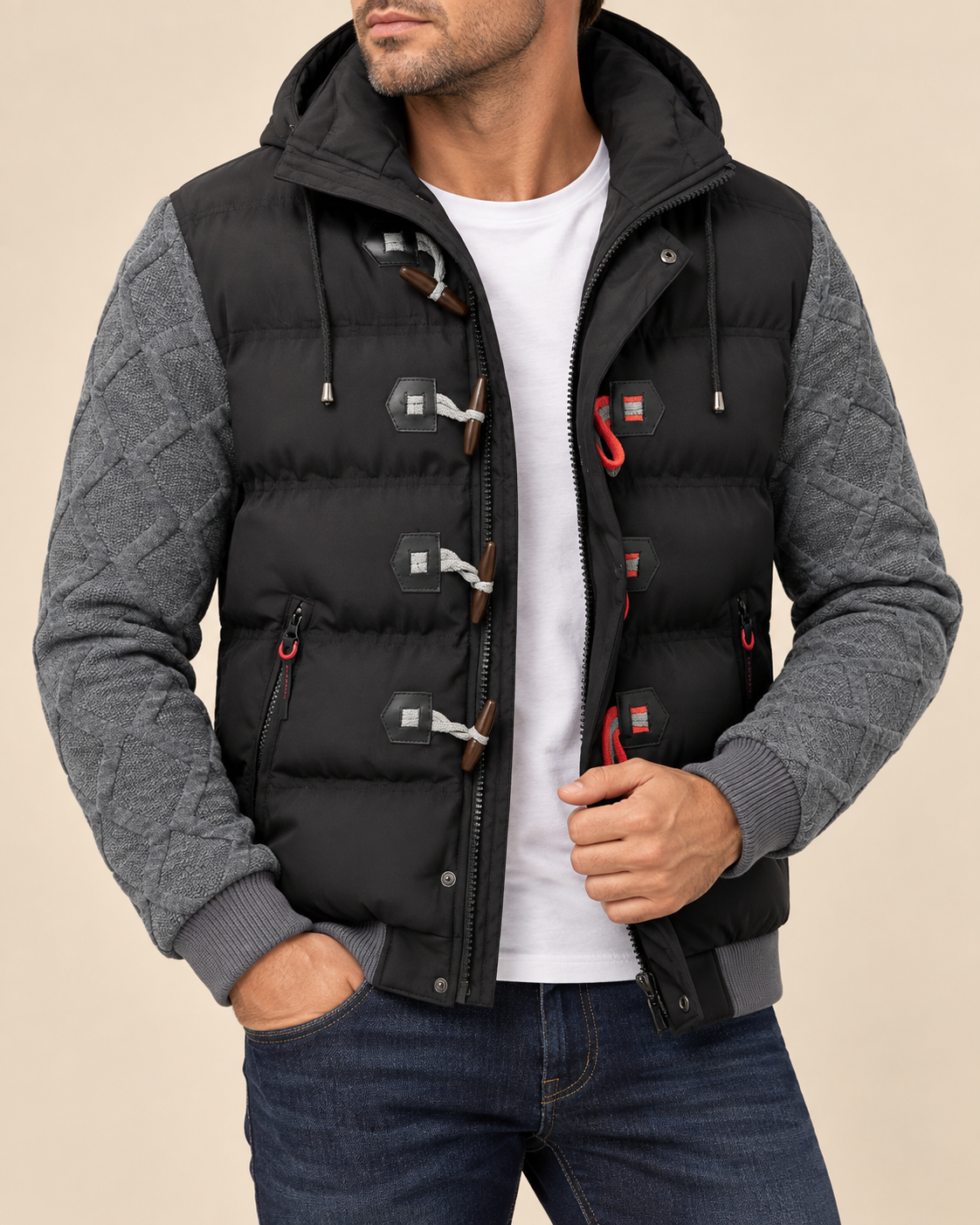 A man wearing a black hooded puffer vest with gray quilted sleeves, toggle closures, and a white t-shirt underneath, paired with blue jeans against a beige background.