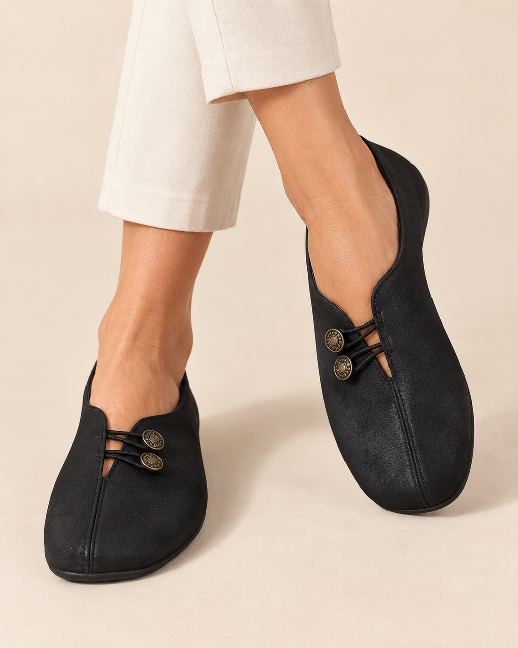 A person wearing black slip-on mule-style shoes with decorative cutouts and two metallic buttons on each shoe, paired with light beige cropped pants against a warm beige studio background.