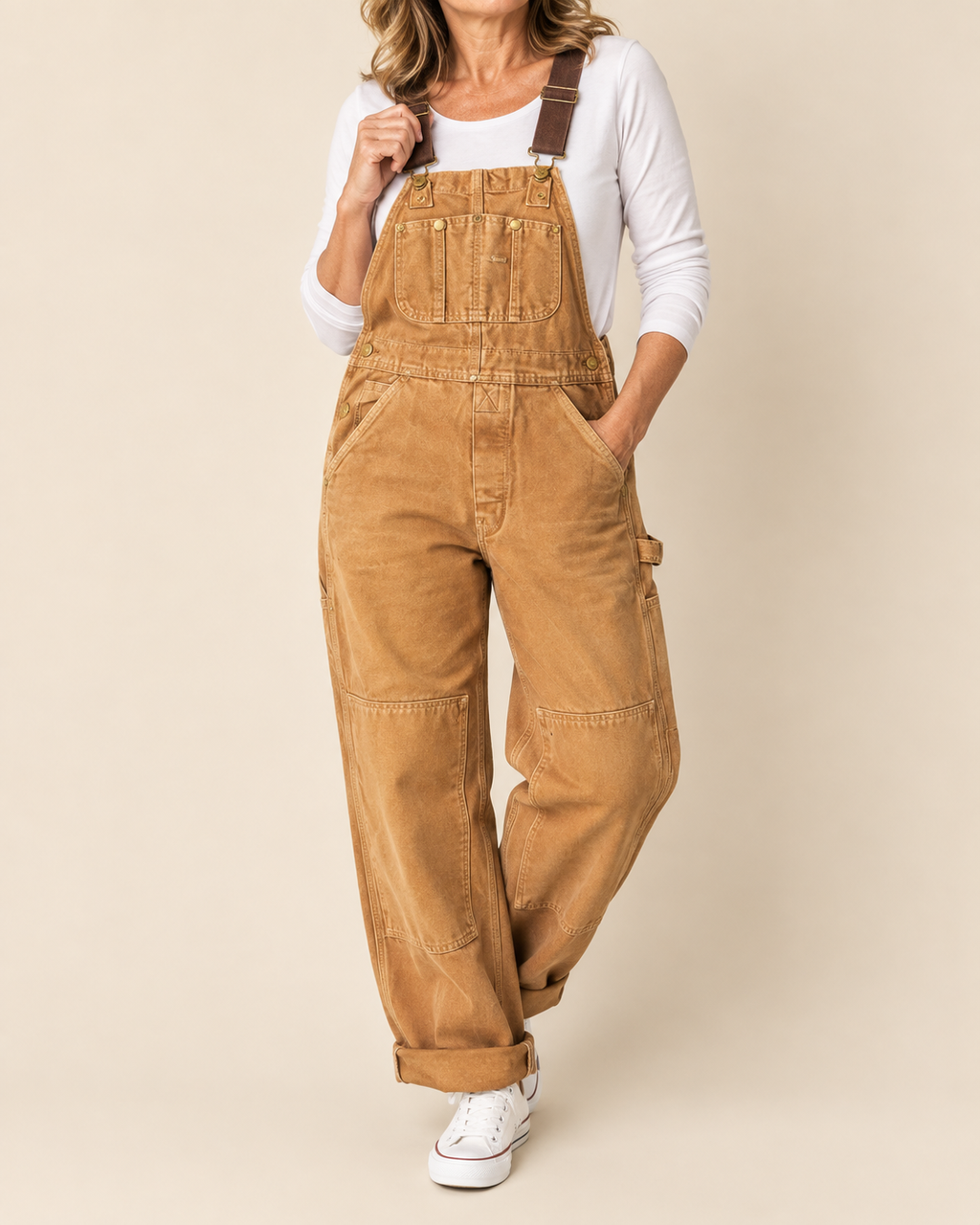 A woman wearing tan corduroy work overalls with brown adjustable shoulder straps, a white long-sleeve top, and white sneakers, standing against a solid beige studio background.