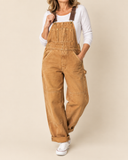 A woman wearing tan corduroy work overalls with brown adjustable shoulder straps, a white long-sleeve top, and white sneakers, standing against a solid beige studio background.