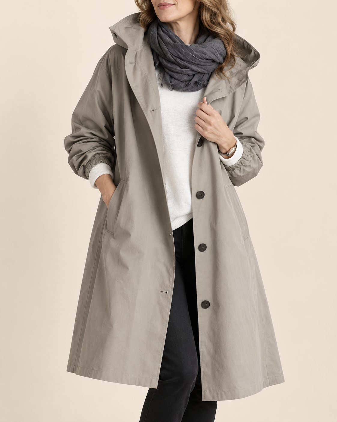 A woman wearing a mid-length light gray hooded raincoat over a white top, black pants, and a dark gray scarf, photographed against a plain beige studio background.
