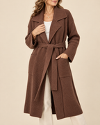 A woman wearing a long brown ribbed knit belted coat over a white top and white pants, photographed against a plain warm beige studio background.