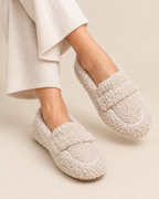 A pair of beige sherpa fleece slip-on loafers worn with cream ribbed knit pants, shown against a solid light beige background.