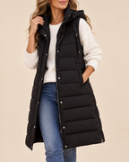 A woman wearing a long black puffer vest over a white knit sweater and blue jeans, standing against a plain beige studio background.