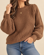 A woman wearing a chunky brown knit sweater with balloon sleeves and blue jeans, shown from the neck down against a plain beige background.