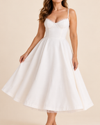 A woman wearing a white A-line midi dress with a sweetheart neckline, structured corset-style bodice, thin spaghetti straps, and a flared skirt, posed against a solid beige studio background.