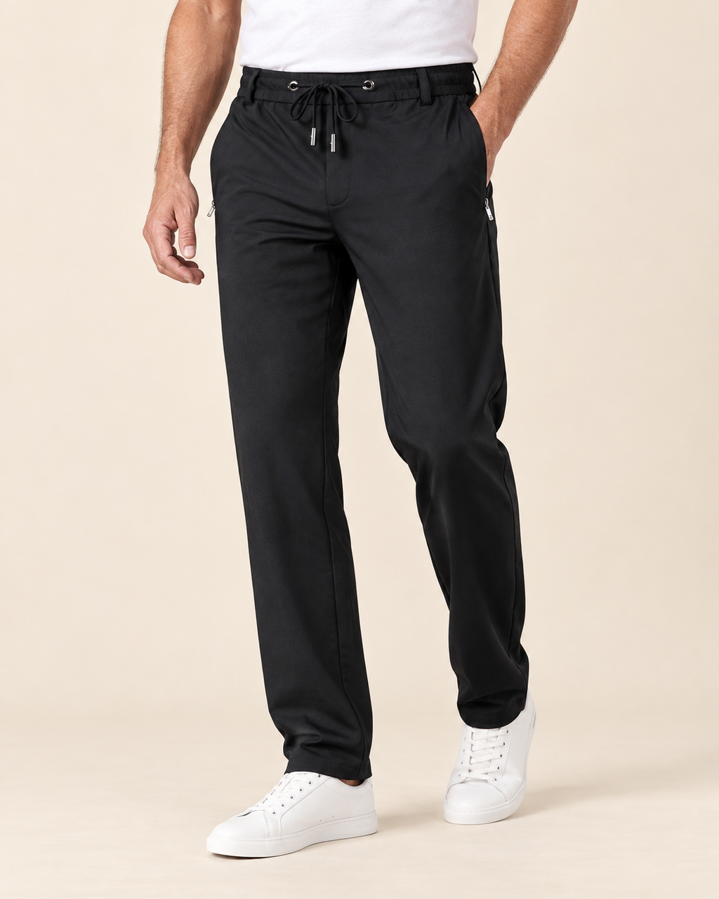 A man wearing black jogger-style pants with a drawstring waist and white sneakers, photographed against a plain warm beige studio background.