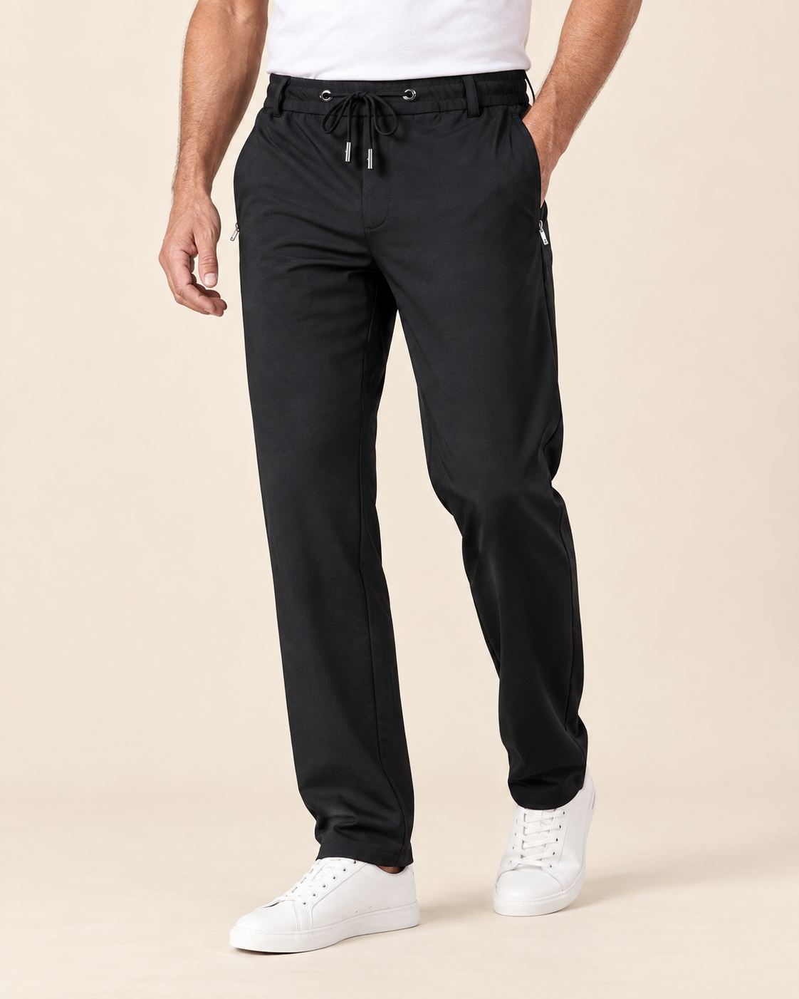 A man wearing black jogger-style pants with a drawstring waist and white sneakers, photographed against a plain warm beige studio background.
