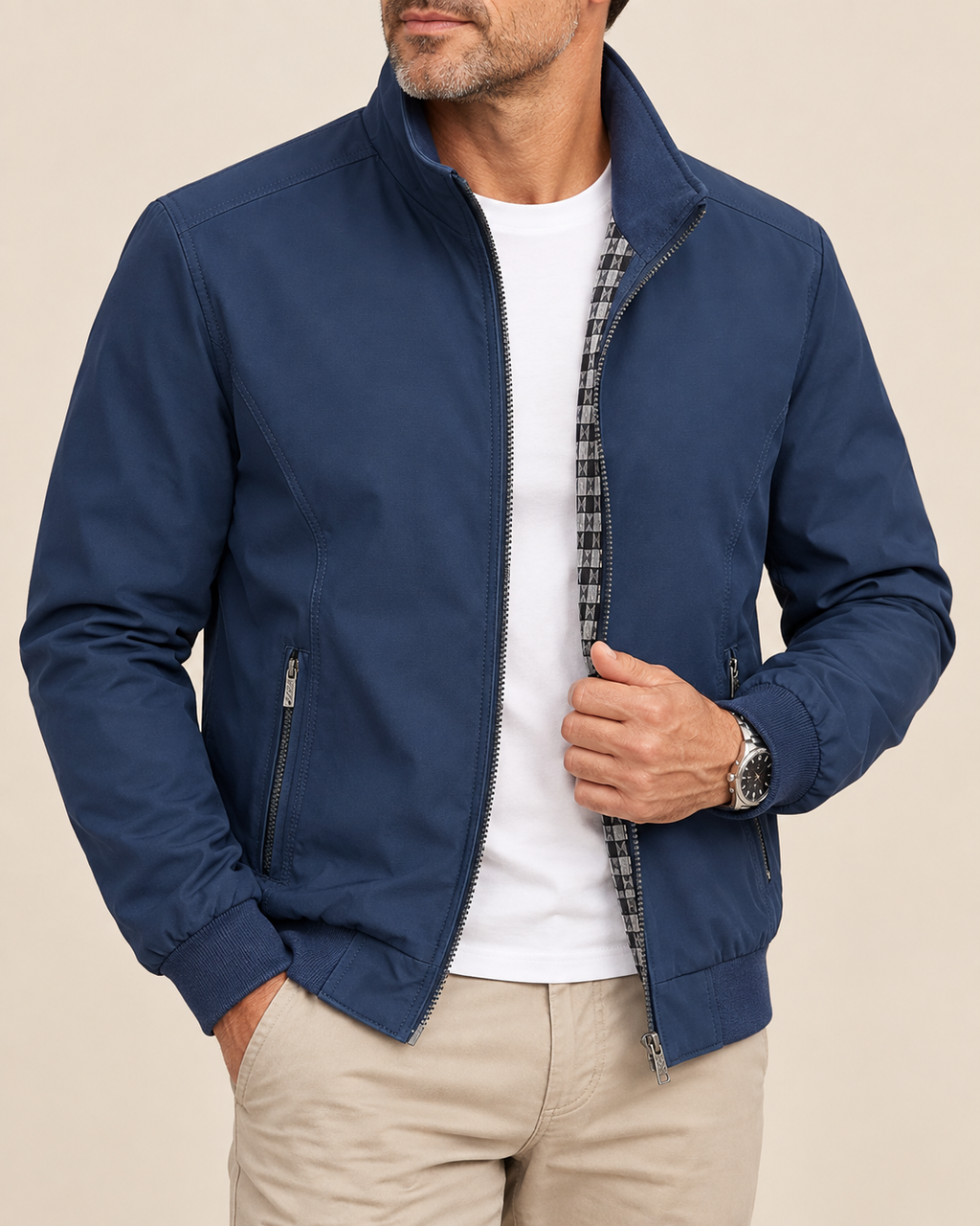 A man wearing a navy blue zip-up jacket with a high collar, styled over a white t-shirt and beige pants, shown from the chin down against a solid beige background.
