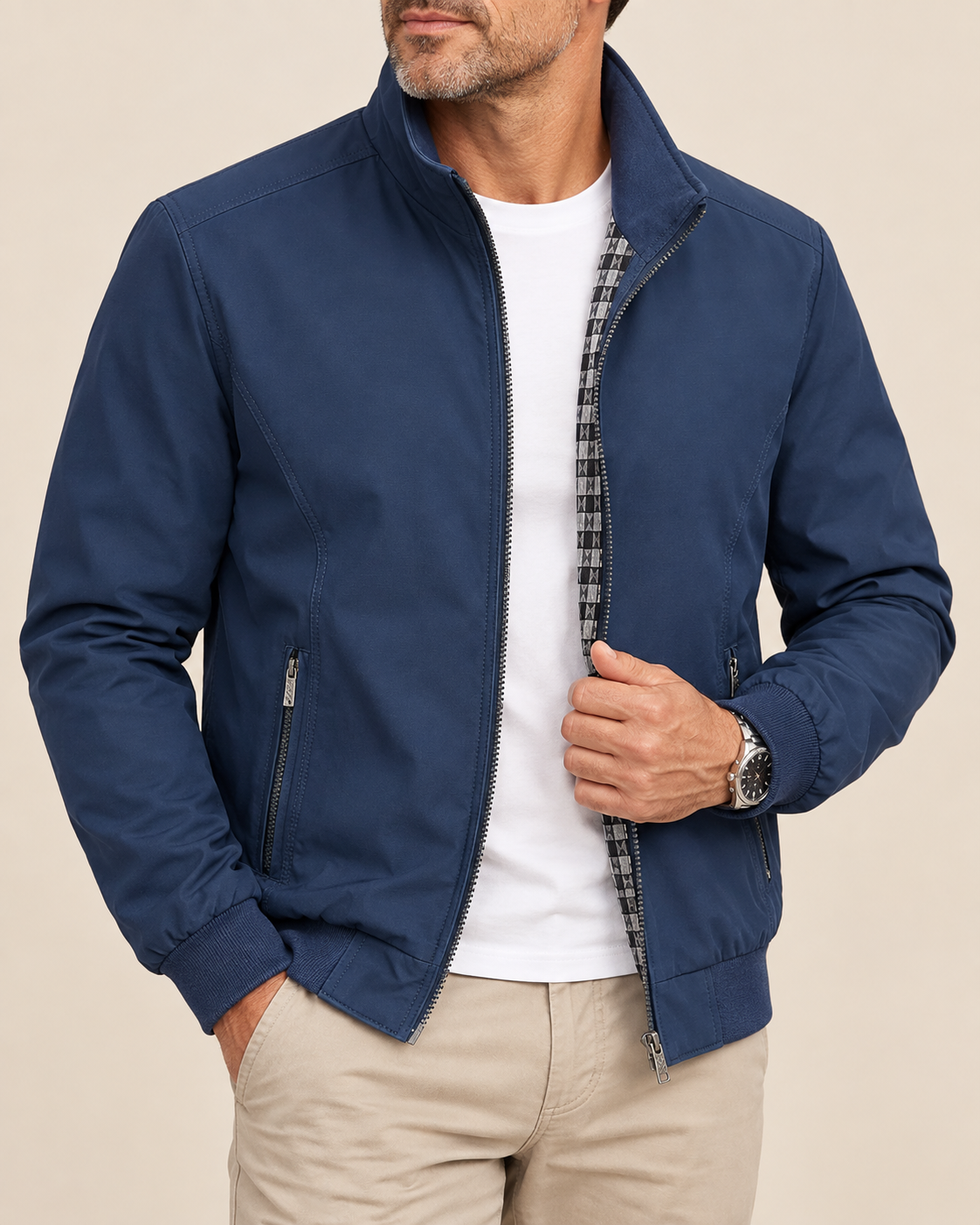 A man wearing a navy blue zip-up jacket with a high collar, styled over a white t-shirt and beige pants, shown from the chin down against a solid beige background.