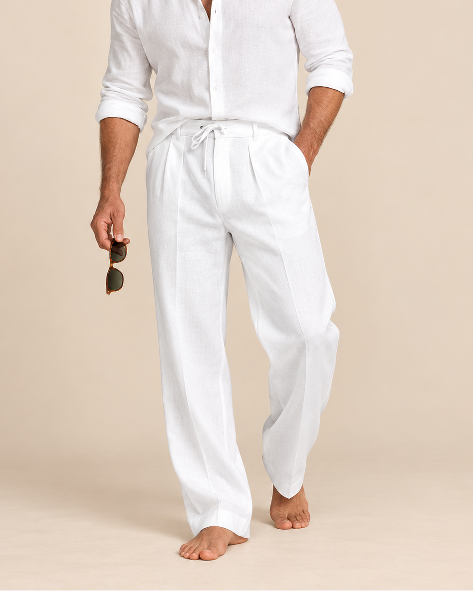 A man wearing a white long-sleeve linen shirt and matching white drawstring trousers, standing barefoot against a warm beige studio background and holding sunglasses.