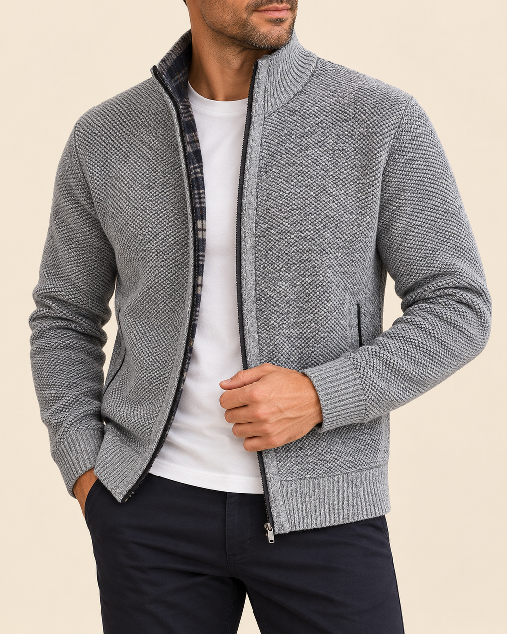 A man wearing a gray textured zip-up knit cardigan over a white t-shirt and dark pants, shown from the chin down against a solid beige background.