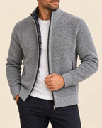 A man wearing a gray textured zip-up knit cardigan over a white t-shirt and dark pants, shown from the chin down against a solid beige background.