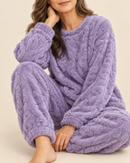 A woman wearing a cozy, textured lavender fleece pajama set with a cable-knit pattern, sitting against a solid beige background.