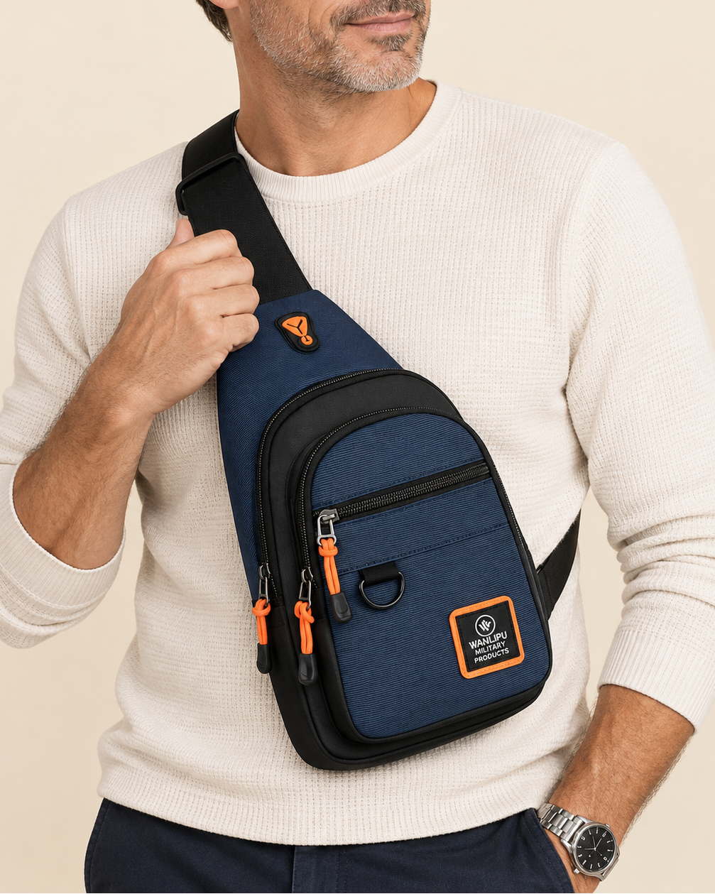 A man wearing a navy blue and black sling bag with orange zipper pulls and a logo patch, styled with a cream ribbed knit sweater against a light beige background.