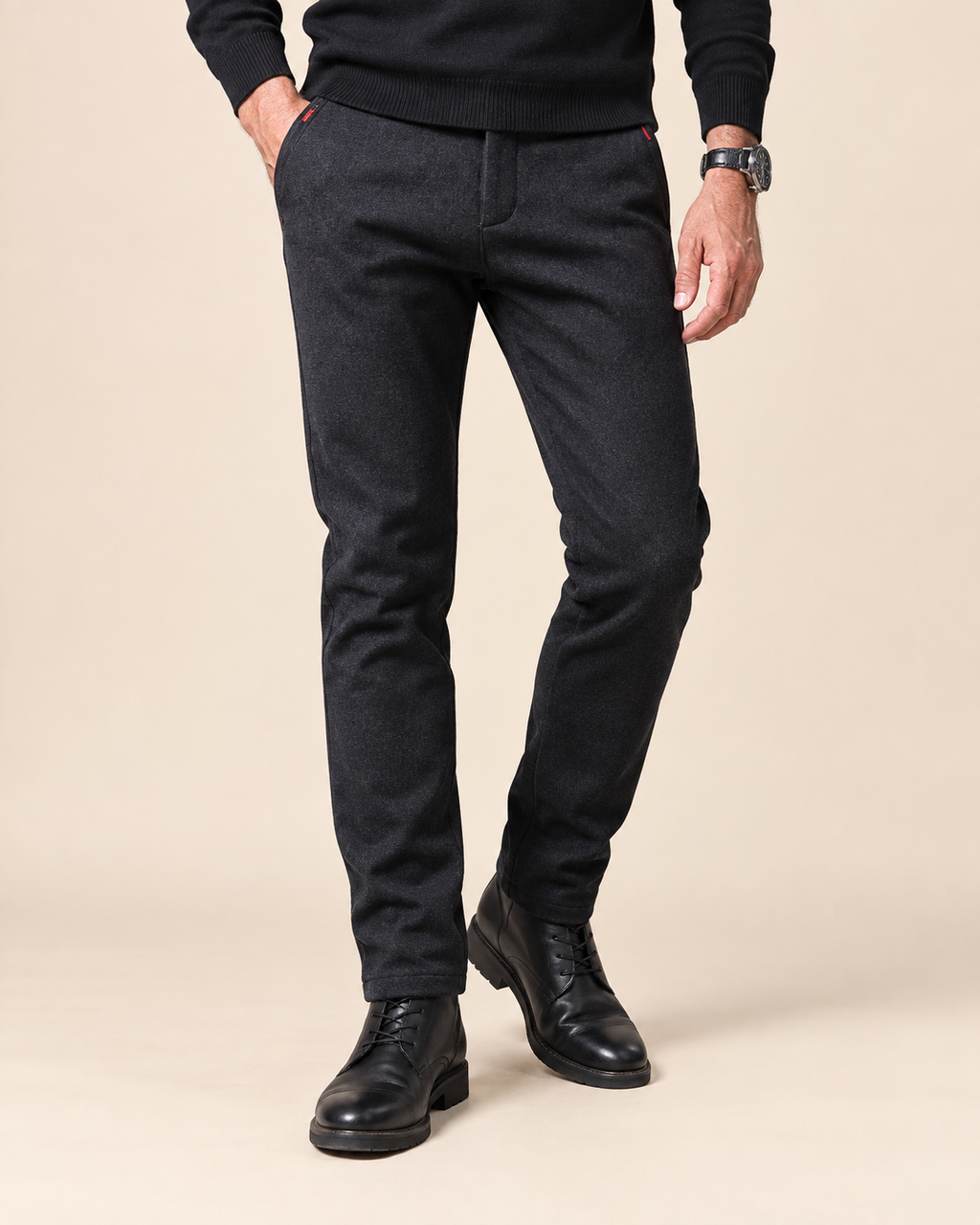 A man wearing slim-fit dark charcoal gray wool trousers, a black sweater, and black leather dress shoes, standing against a solid beige studio background.