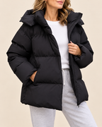 A woman wearing a black oversized puffer jacket with a high padded collar, zippered front, and side pockets, styled over a white top and light gray sweatpants against a plain beige studio background.