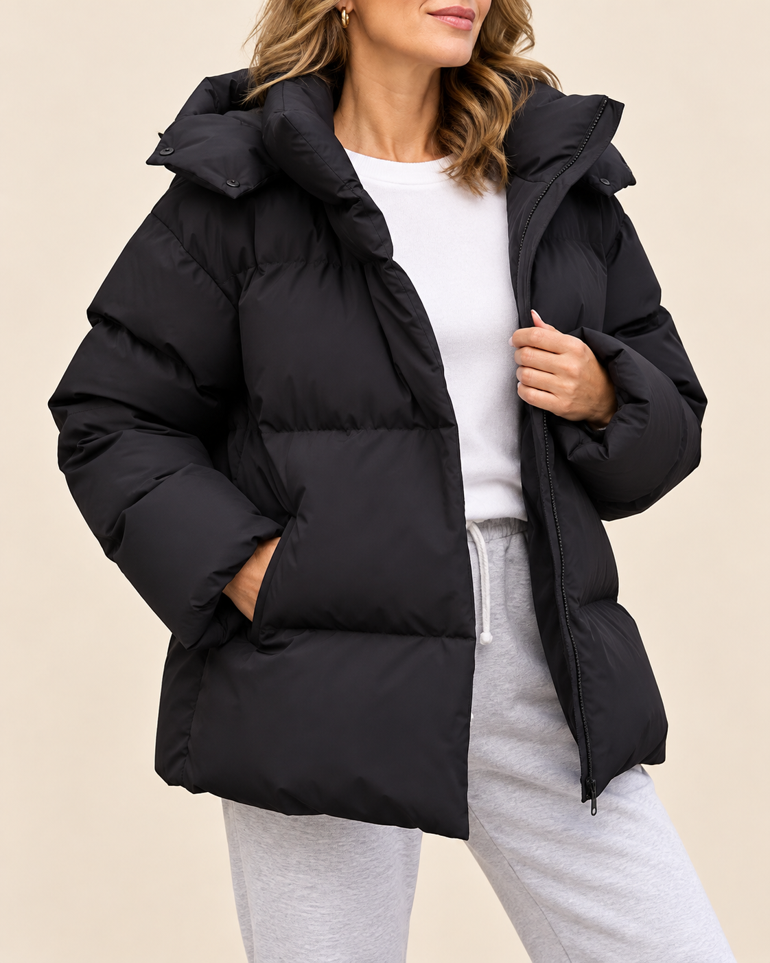 A woman wearing a black oversized puffer jacket with a high padded collar, zippered front, and side pockets, styled over a white top and light gray sweatpants against a plain beige studio background.