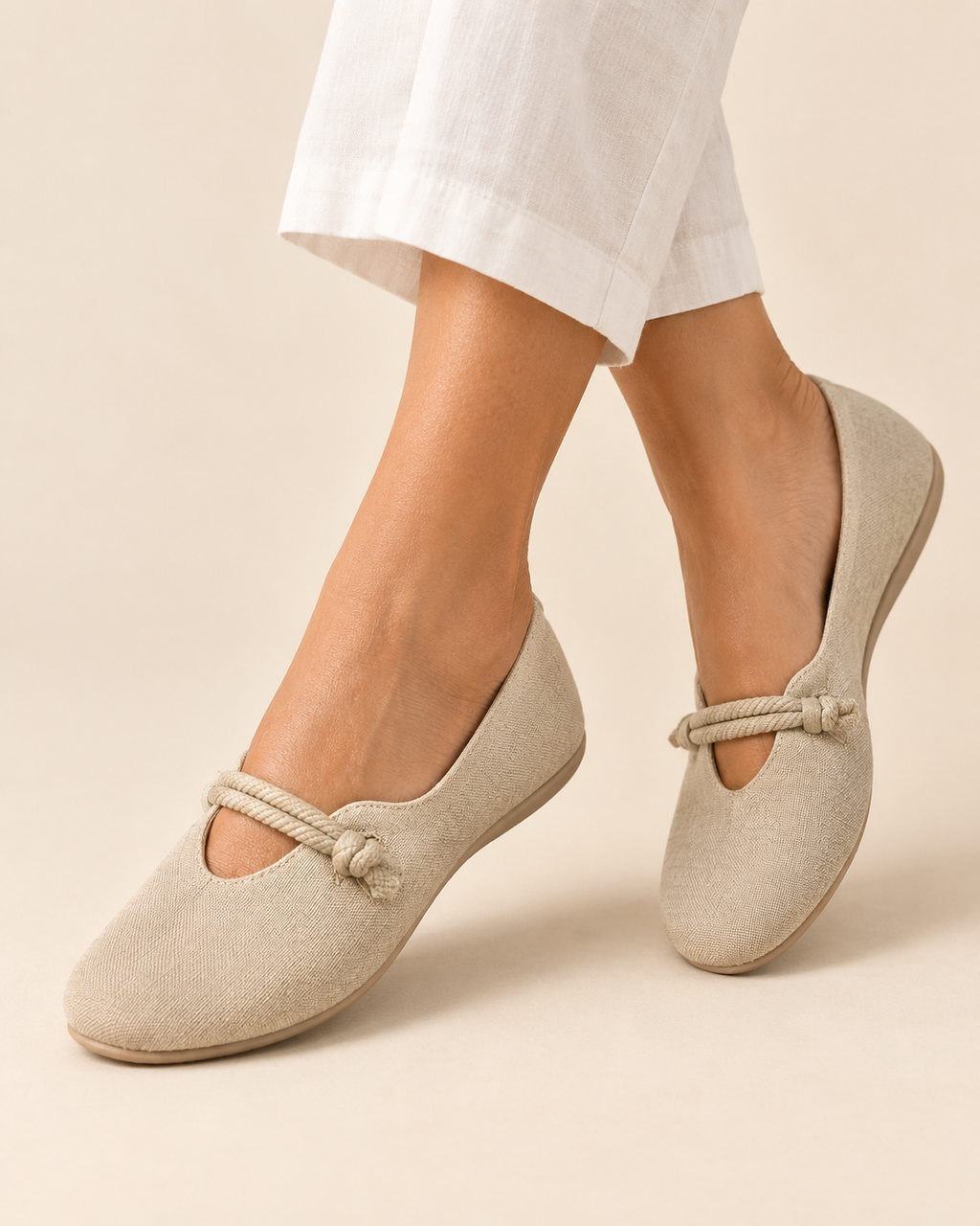A person wearing light beige Mary Jane-style flats with a rope detail across the top, shown against a plain warm beige studio background. White pants are visible above the feet.
