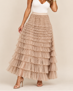 A woman modeling a long, tiered tulle maxi skirt in a light taupe-beige color, paired with a white ribbed tank top and nude high-heeled sandals, against a solid beige studio background.